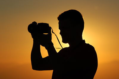 Silhouette man photographing at camera against sky during sunset