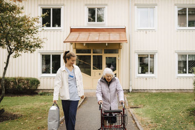 Elderly woman walking with walker by female caregiver in front yard