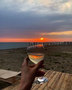 Hand holding drink at beach against sky during sunset