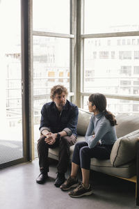 Full length of mald professional talking to businesswoman while sitting on sofa at office