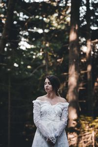 Thoughtful young bride standing against trees in forest
