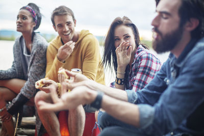 Happy friends holding hotdogs while sitting by river