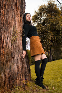 Low angle portrait of young woman leaning on tree while standing on field at park