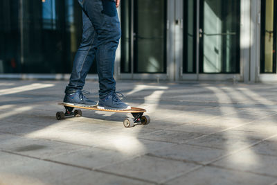 Hipster in dark blue jeans and sneakers riding on longboard in motion on asphalt.