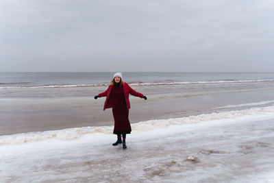 A joyful woman in a red jacket and a dress by the sea in winter