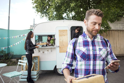 Male customer using smart phone on street with food truck owners and woman in background