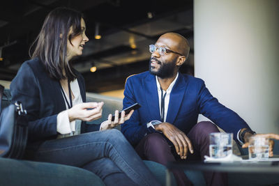 Businesswoman with smart phone talking to colleague while sitting at workplace