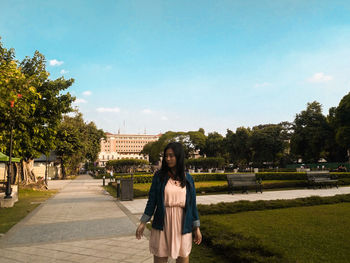 Young woman looking away while standing in park against blue sky