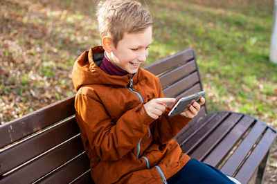 The boy is playing on the phone sitting on a park bench in autumn and smiling