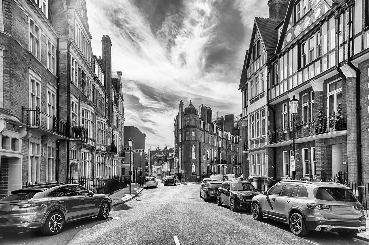 architecture, car, mode of transportation, motor vehicle, road, transportation, city, building exterior, black and white, street, built structure, monochrome, infrastructure, monochrome photography, lane, land vehicle, urban area, neighbourhood, cityscape, building, residential area, sky, cloud, city street, vehicle, nature, residential district, city life, day, metropolis, traffic, travel, motion, the way forward, alley, no people, outdoors, travel destinations, house