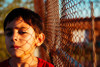 Portrait of boy standing with chain link fence shadow on face outdoors
