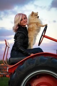 Portrait of smiling young woman with dog sitting on tractor at sunset