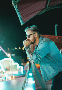 Side view of man wearing sunglasses standing by railing at night