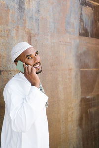 Cheerful muslim male in traditional clothes smiling and browsing cellphone while standing near shabby wall on street