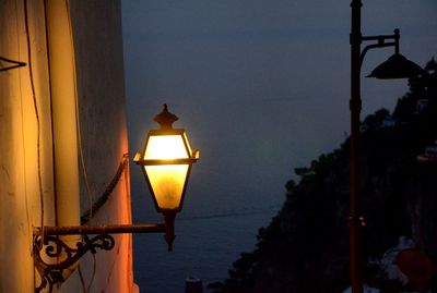 Illuminated street light by sea against sky during sunset