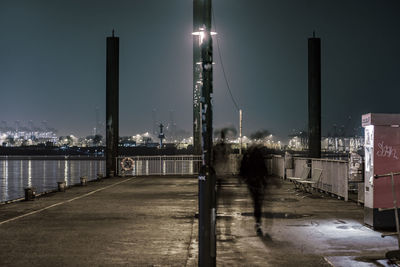 Illuminated street by river against sky at night