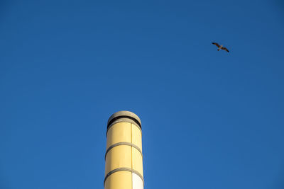 Low angle view of bird flying over smoke stack against clear blue sky