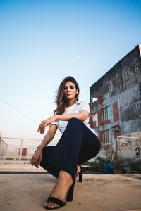 Low angle portrait of young woman crouching against clear sky