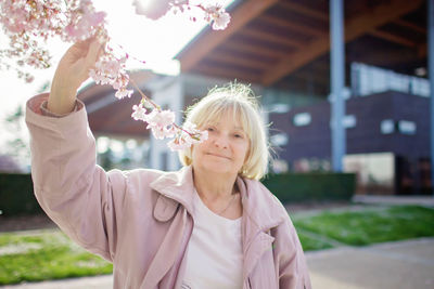 Senior woman in pink coat smells blooming branch of sakura during her walking in the spring garden