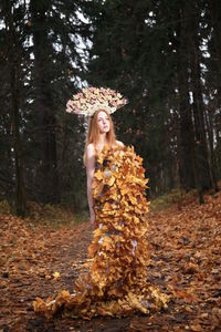 Thoughtful young woman covered with leaves standing at forest during autumn