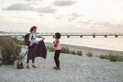 Girl looking at woman holding towel at beach against sky during sunset