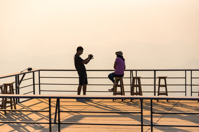 Friends standing on railing against sky during sunset