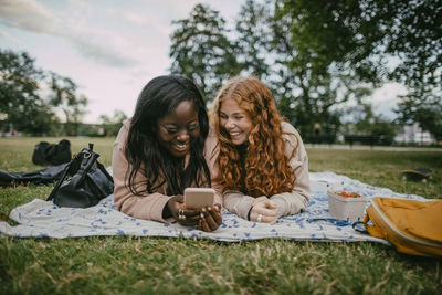 Happy multiracial female friends using smart phone while lying on picnic blanket at park