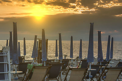 Deck chairs and closed umbrellas at beach against cloudy sky during sunset