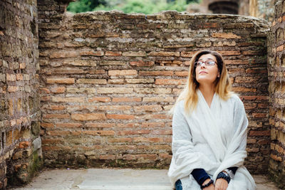 Young woman looking away while sitting against brick wall