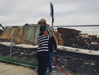 Rear view of men standing by chainlink fence against sky