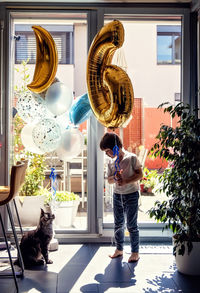 Toddler boy holding balloon in shape of figure six looking at his maine coon cat friend at home