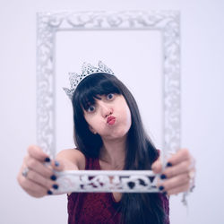 Portrait of young woman wearing crown holding picture frame while standing against white background