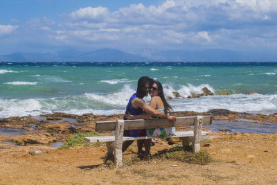 Portrait of two woman sitting on bench at beach against sky