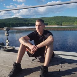 Portrait of young man sitting on shore against sky