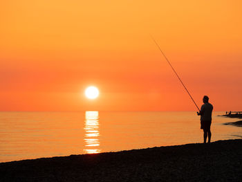 Silhouette man fishing in sea against orange sky