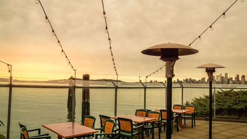 Empty chairs and table by sea against sky during sunset