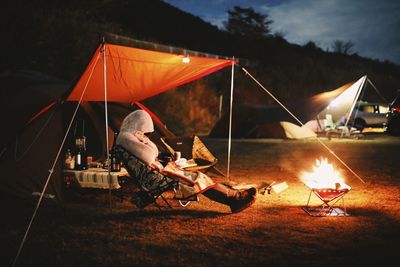 Woman sitting by fire pit in tent against sky at night