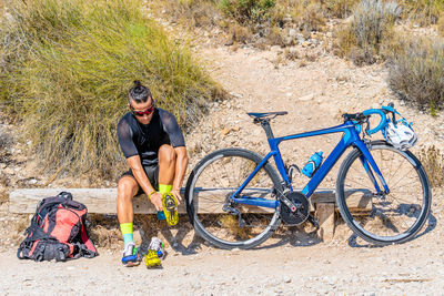 Full length of active male bicyclist in sportswear sitting on wooden bench and putting on cycling shoes while preparing for mountain bike ride