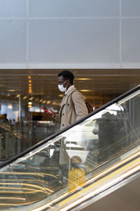 Black traveler man stands on escalator in airport wear face mask. covid-19, transportation concept.