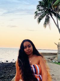 Portrait of young woman sitting at beach against sky during sunset