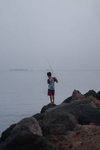 Rear view of boy fishing in sea against sky