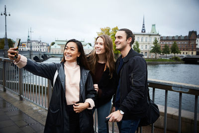 Smiling woman taking selfie through mobile phone while standing on bridge in city