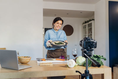 Smiling vlogger holding food plate filming through camera at home