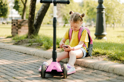 A smiling little schoolgirl girl is sitting on the curb with a phone in her hands during a break