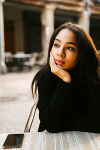 Young woman in black jumper leaning on hand and looking in thoughts away resting at sidewalk cafe table