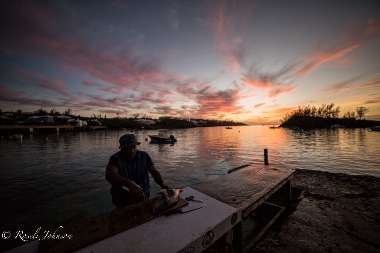 MAN SITTING AT LAKE AGAINST SKY DURING SUNSET