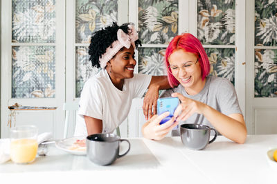 Young woman with pink hair smiling and showing smartphone to cheerful african american friend while having breakfast at home together