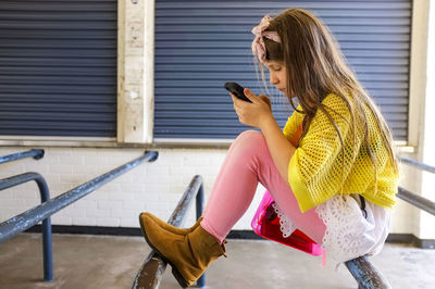 Side view of girl using mobile phone while sitting on pole