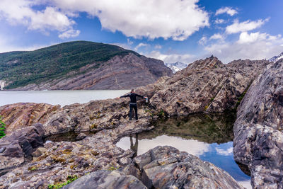 Rear view of woman standing on rock by lake and pond against mountains and sky