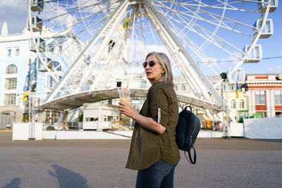 Stylish, confident mature woman with grey hair enjoys refreshing juice cup in vibrant european city.